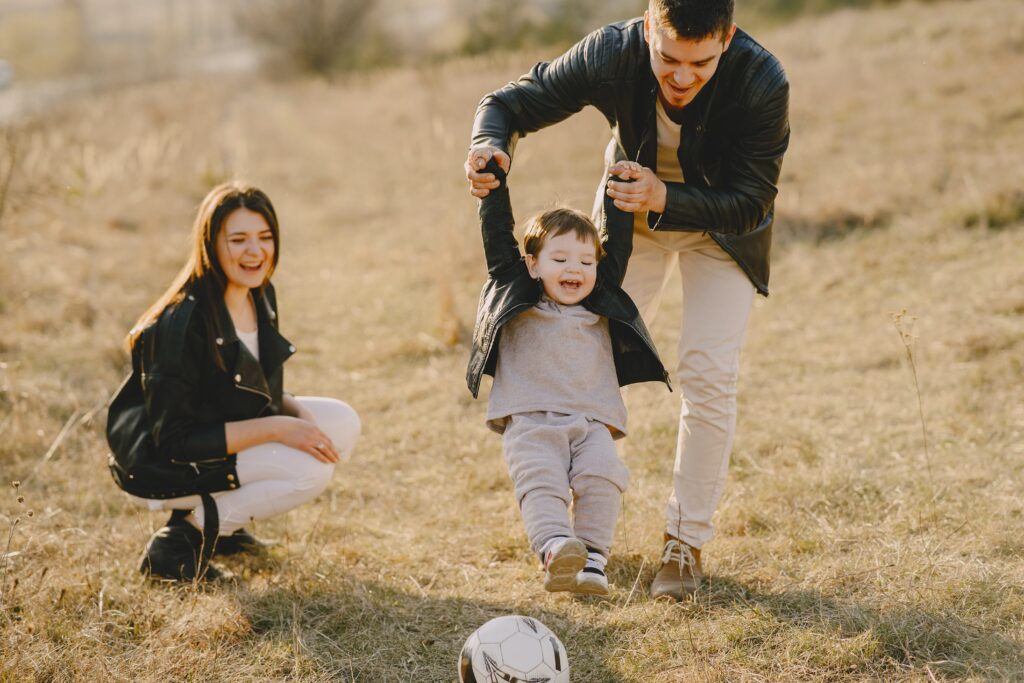 father holding child's hands to play soccer while mother crouches down and watches